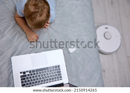 Little blond boy in white wireless headphones uses a modern laptop on the bed, gets new knowledge using a portable device while a robot vacuum cleaner does the cleaning. Top view