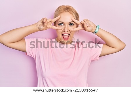 Young blonde woman wearing casual pink t shirt doing peace symbol with fingers over face, smiling cheerful showing victory 