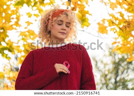 Breast cancer survivor. Outdoor shot of a peaceful caucasian young adult woman holding pink ribbon in front of her chest. Autumn park vibes. High quality photo