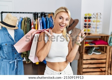 Young caucasian woman holding shopping bags at retail shop with a big smile on face, pointing with hand finger to the side looking at the camera. 