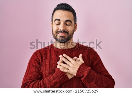 Young hispanic man with beard wearing casual sweater over pink background smiling with hands on chest with closed eyes and grateful gesture on face. health concept. 