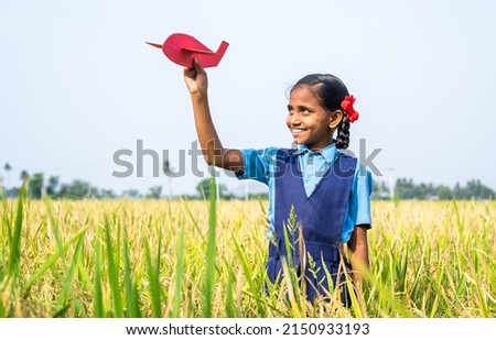 Happy village girl kid with school uniform playing using toy cardboard aeroplane at paddy field - concept of childhood dream, aspirations and freedom. Royalty-Free Stock Photo #2150933193