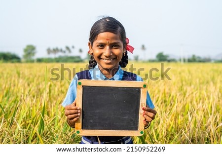 Happy smiling school girl kid holding empty slate board near paddy field - concept of education, learning and childhood empowerment