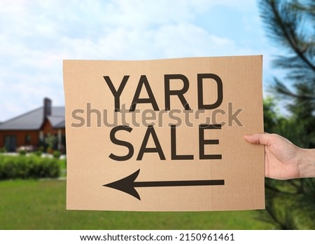 Woman holding sign with text YARD SALE and blurred view of modern house on sunny day