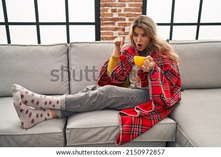 Young woman sitting on the sofa drinking a coffee at home pointing down with fingers showing advertisement, surprised face and open mouth 