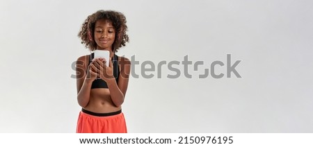 Smiling athletic black girl wearing earphones watching something on smartphone. Curly female child wearing sportswear. Modern youngster lifestyle. Isolated on white background in studio. Copy space