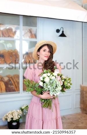 young beautiful happy girl wearing a pink dress, straw hat, holding a bouquet of flowers, posing in the street of a European city near a coffee shop. Spring, summer fashion. Copy space