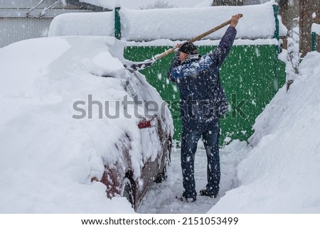 A man is shoveling snow off a red car. Large flakes of snow falling. Nearby are large drifts of snow Royalty-Free Stock Photo #2151053499