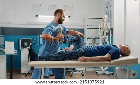 Therapist using legs raise exercise to stretch muscles and crack bones, helping senior man with mechanical disorders in cabinet. Male osteopath doing alternative medicine procedure. Royalty-Free Stock Photo #2151075921