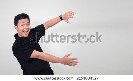 Portrait of excited Asian man in black polo shirt smiling and looking at the camera pointing and spreading his hands to the side. Isolated image on gray background