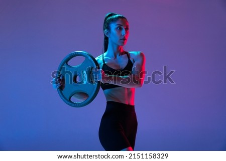 One muscled woman in sportswear training with disk of barbell isolated on purple background in neon light. Sport, beauty, strength concept. Female sports