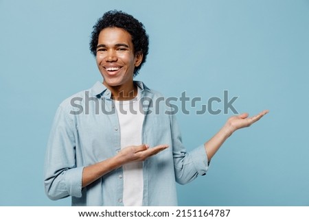 Overjoyed happy fun young black curly man 20s years old wears white shirt pointing palms hands aside on workspace area copy space mock up isolated on plain pastel light blue background studio portrait