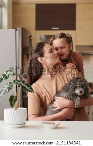Girl telling something to mother sitting at kitchen table with small dog in hands
