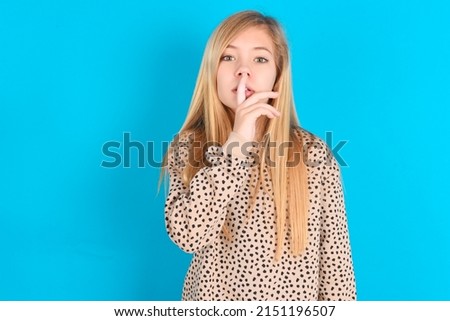 little caucasian kid girl wearing animal print sweater over blue background  makes silence gesture, keeps finger over lips. Silence and secret concept.