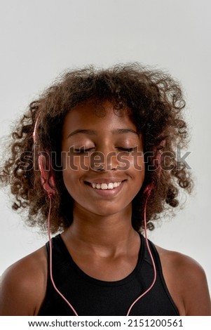 Portrait of smiling sports black girl with closed eyes listening music in earphones and enjoying it. Curly female child wearing tank top. Modern youngster lifestyle. White background. Studio shoot