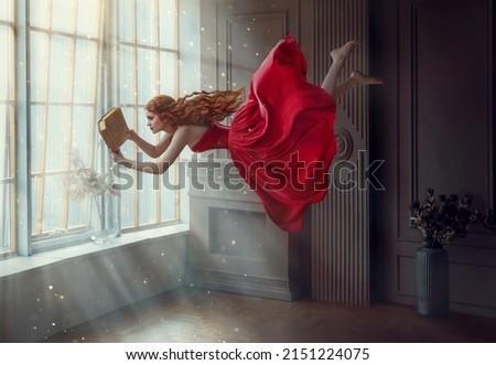 Fantasy redhead woman soars floats flies in air. Art photo levitation. Girl fairy princess reads magic book, divine light from window. Red midi dress, lon hair flutters in wind. Room classic interior Royalty-Free Stock Photo #2151224075