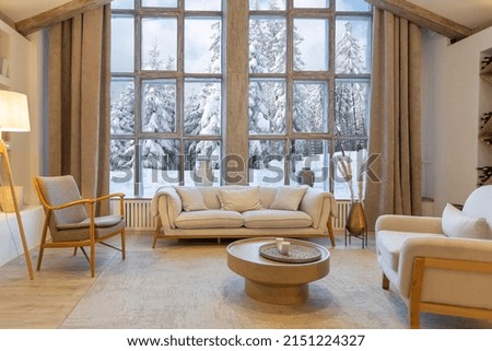 cozy warm home interior of a chic country chalet with a huge panoramic window overlooking the winter forest. open plan, wood decoration, warm colors and a family hearth Royalty-Free Stock Photo #2151224327