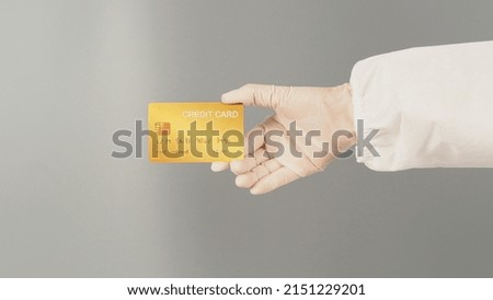 Hand is holding gold credit card on grey background. Hand wears a PPE suit and a white medical glove.