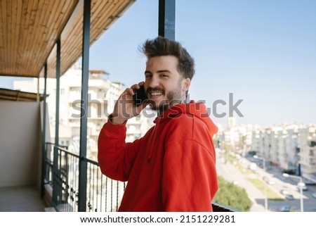 Smiling young man wearing red hoodie talking by smartphone while leaning metal fences looking at the camera with big smiles. Talking with girlfriend.