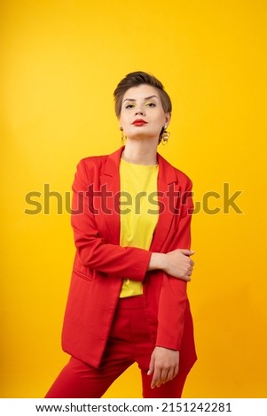 A woman in bright makeup, a red jacket, a yellow T-shirt and red trousers, with bright red lipstick and green shadows, stands on a yellow background with arm hold another elbow. Studio shooting