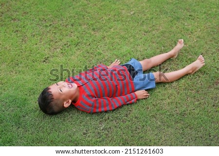 Happy Asian little boy lying down on green lawn. Kid lies on grass looking on sky. Royalty-Free Stock Photo #2151261603