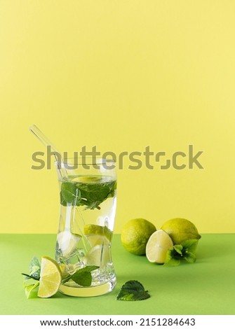 Mojito cocktail with lime and mint in a tall transparent glass on a green table. Lime wedges and mint leaves in the background. Yellow background, text space