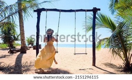 Slow motion of unrecognizable travel woman on wooden swing on tropical sandy beach with blue sea and palm trees. Female traveler on Phuket island, Thailand. Vacation and summer holidays concept.  Royalty-Free Stock Photo #2151296115