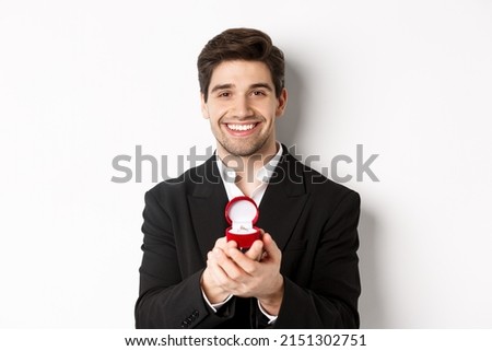 Image of handsome man looking romantic, open small box with engagement ring, making a proposal and smiling, standing against white background