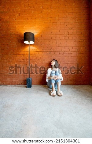 Young woman sitting alone on floor in empty room with floor lamp on brick wall background. Concept of loneliness and solitude at home Royalty-Free Stock Photo #2151304301