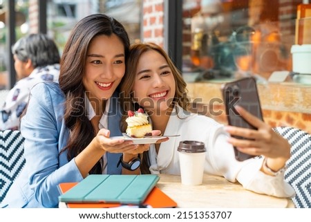 Asian woman friends using smartphone selfie together while sitting at outdoor coffee shop eating bakery and drinking coffee in urban city street. Beautiful female enjoy outdoor lifestyle in the city