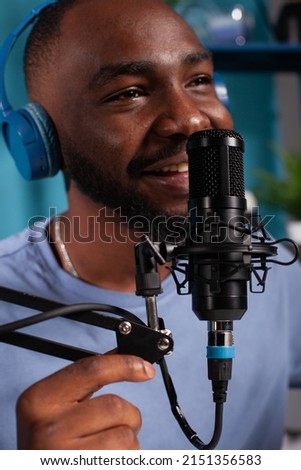 Closeup of smiling vlogger talking to audience using professional microphone wearing wireless headphones in live podcast. Close focus on studio mic used by content creator in online interview.