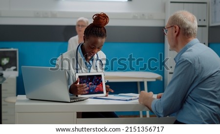 Black doctor holding virus animation on tablet showing disease cells to elder patient at desk. African american medic explaining coronavirus bacteria using modern technology on device.