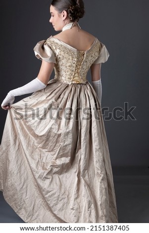 A Victorian woman wearing a gold ball gown with long, fingerless lace gloves and standing against a studio backdrop Royalty-Free Stock Photo #2151387405