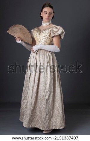 A Victorian woman wearing a gold ball gown with long, fingerless lace gloves and standing against a studio backdrop Royalty-Free Stock Photo #2151387407