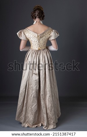 A Victorian woman wearing a gold ball gown with long, fingerless lace gloves and standing against a studio backdrop Royalty-Free Stock Photo #2151387417