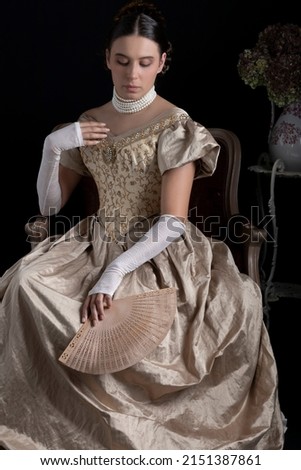 A Victorian woman in a gold ball dress sitting on an ornate chair against a dark backdrop Royalty-Free Stock Photo #2151387861