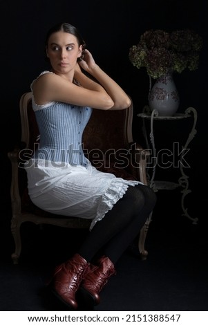 A Victorian woman wearing a corset and drawers and sitting on an ornate chair in a darkened room