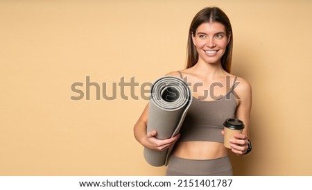 Beautiful smiling healthy female fitness trainer in stylish brown crop top and leggings holding rolled mat and plastic cup of coffee after workout classes over peach color background