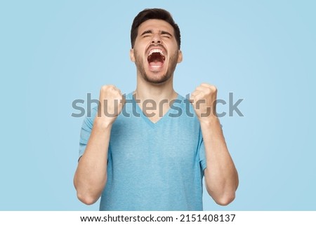 Young man shouting with closed eyes, celebrating victory, squeezing fists in deep emotional expression of happiness and luck, isolated on blue background  Royalty-Free Stock Photo #2151408137