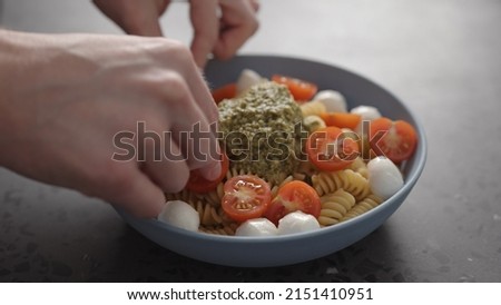 man add cherry tomatoes to pesto fusilli pasta in blue bowl on concrete background wide photo