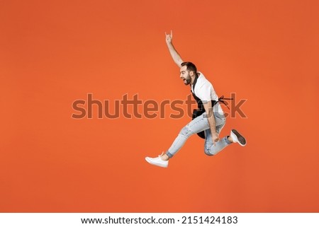 Full length young man barista bartender barman in apron white t-shirt work in coffee shop jump horns up gesture, depicting heavy metal rock sign isolated on orange background. Small business startup