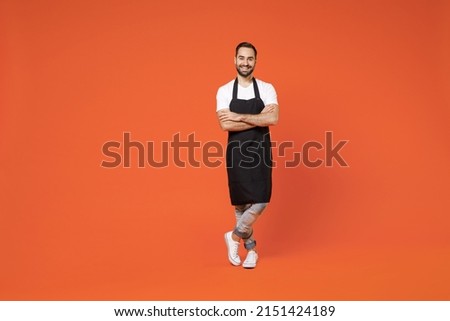 Full length young man barista bartender barman employee in black apron white t-shirt work in coffee shop hold hands crossed folded isolated on orange background studio. Small business startup concept Royalty-Free Stock Photo #2151424189