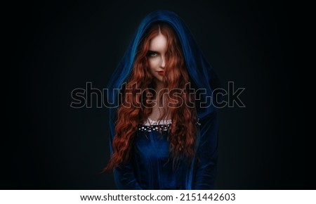 Portrait fantasy gothic red-haired woman witch. Vampire girl in blue medieval dress, vintage old historical style hood on head. Black background. Red lips, hair flying soar in wind. halloween costume Royalty-Free Stock Photo #2151442603
