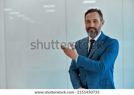 Waist up of happy bearded man in suit pointing at white wall by hand, copy space Royalty-Free Stock Photo #2151450733