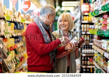 Cheerful stylish senior couple husband and wife making shopping together at supermarket, walking by stire aisle full of goods, choosing pasta sauce and having conversation, smiling Royalty-Free Stock Photo #2151466439