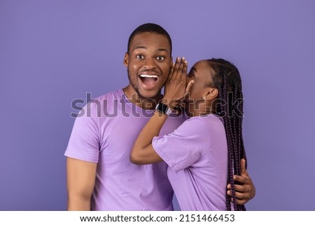 Smiling black lady sharing secret or whispering gossips into her boyfriend's ear on violet purple studio background. Millennial African American couple sharing shocking news, discussing rumors Royalty-Free Stock Photo #2151466453