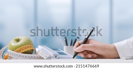 Professional nutritionist sitting at desk and writing a meal plan, measuring tape and apple Royalty-Free Stock Photo #2151476669