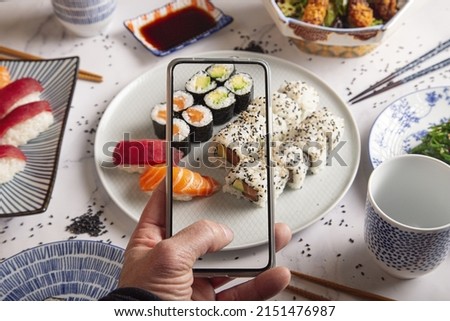 A young man taking a photo of a plate with Japanese sushi nigiri and rolls on a smartphone