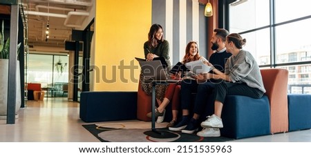 Multicultural businesspeople working in an office lobby. Group of happy businesspeople smiling while sitting together in a co-working space. Young entrepreneurs collaborating on a new project. Royalty-Free Stock Photo #2151535963