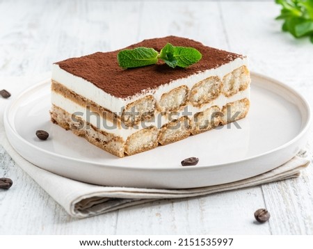 Tiramisu cake decorated with cocoa powder and fresh green mint leaf on white ceramic plate. Close up food. Traditional italian dessert. Coffee beans, textile napkin, white wooden table background. Royalty-Free Stock Photo #2151535997